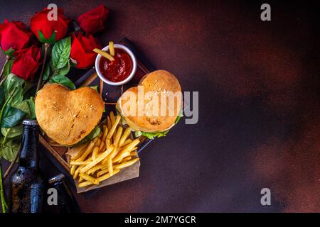 Herzförmige Burger zum Valentinstag. Zwei leckere Cheeseburger mit pommes Frites und Bierflaschen auf dunklem Tischhintergrund. Eine Idee für das Valentinstag-Dinner Stockfoto