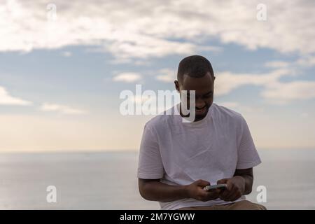 Glücklicher junger afroamerikanischer Mann in lässiger Kleidung, der in der Nähe des Meeres gegen den bewölkten Himmel bei Sonnenuntergang auf seinem Handy surft Stockfoto