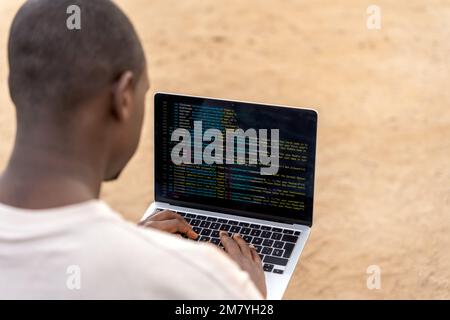 Ein Afroamerikaner in lässiger Kleidung, der seinen Laptop benutzt, um Code für Software zu erstellen, während er Remote am Strand arbeitet Stockfoto