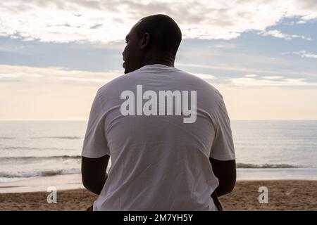 Rückansicht eines afroamerikanischen Mannes in weißer Freizeitkleidung, der abblickt, während er bei Sonnenuntergang am Sandstrand gegen den bewölkten Himmel sitzt Stockfoto