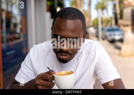 Konzentrierter junger bärtiger afroamerikanischer Mann in weißem T-Shirt, der heißen Kaffee im Café im Freien in der Nähe von Palmen trinkt Stockfoto