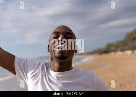 Fröhlicher afroamerikanischer Mann in lässiger Kleidung, lachend und genießt Sommerurlaub am Strand vor bewölktem Himmel Stockfoto