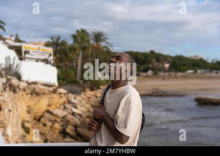 Glücklicher afroamerikanischer Tourist in einem weißen T-Shirt mit Rucksack, der über einen lustigen Witz lacht, während er auf der Terrasse neben dem winkenden Meer steht Stockfoto