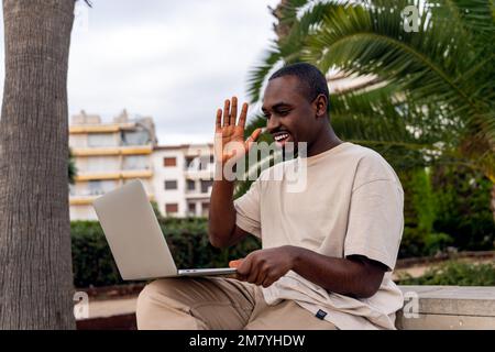 Fröhlicher afroamerikanischer Mann in lässiger Kleidung, der während eines Videoanrufs mit dem Laptop im Park mit der Hand winkt Stockfoto