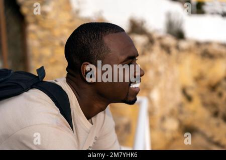 Positiver schwarzer Reisender in lässiger Kleidung mit Rucksack, der sich am Geländer lehnt und wegschaut, während er Zeit am Aussichtspunkt am Strand verbringt Stockfoto