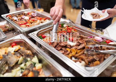 Nahaufnahme eines Mannes, der Teller und Zange hält, während er Essen am Büfetttisch im Hotel oder Restaurant sammelt Stockfoto