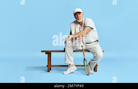 Porträt eines gutaussehenden, seriösen Seniorenmannes in elegantem, weißem Outfit, der auf einer Bank mit Tennisschläger auf blauem Hintergrund sitzt. Freizeitkonzept Stockfoto