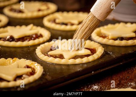 Nahaufnahme von mit Sternen gekrönten Hackkuchen in einer dunklen Metallbrötchenform, die vor dem Backen mit Ei-Waschmittel gebürstet wird Stockfoto