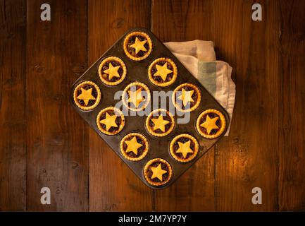 Frisch gebackene Hackkuchen mit Sternen in einer Metallbrötchenform auf dunklem Holzhintergrund: Weihnachtliches Heim, flach gebacken. Stockfoto