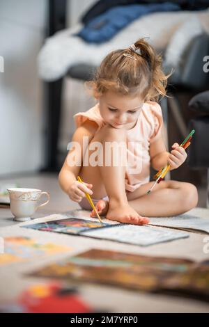 Das kleine Mädchen sitzt zu Hause auf dem Boden und malt mit Farben und Pinseln in einem Malbuch. Frühkindliche Kreativität und Bildung Stockfoto