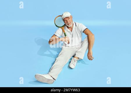 Entspannung. Porträt eines gutaussehenden Seniorenmannes in elegantem, weißem Outfit, der mit Tennisschläger auf blauem Hintergrund posiert. Freizeitkonzept Stockfoto