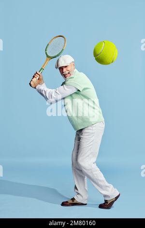 Glücklich. Porträt eines gutaussehenden Seniorenmannes in stilvollem Outfit, der Tennis spielt und Ball auf blauem Hintergrund serviert. Konzept der Freizeitaktivität, Hobby Stockfoto