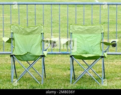 Ansicht der Rückseite von zwei grünen klappbaren Campingstühlen, die vor einer Metallbarriere platziert sind Stockfoto