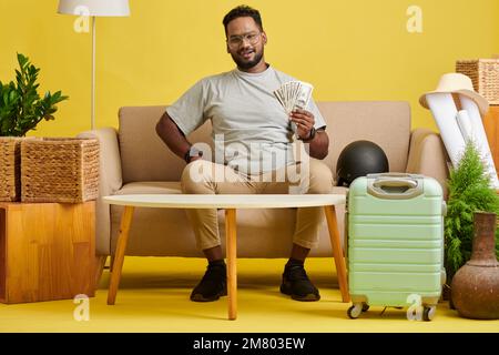 Positiver Mann, der Geld zeigte, das für den Urlaub gespart wurde, als er auf der Couch neben dem Koffer saß Stockfoto