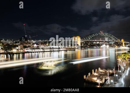 Leichte Wanderwege bei Nacht von Fähren, die am Circular Quay in Sydney ankommen und abfahren, mit Sydney Harbour Bridge im Hintergrund Stockfoto