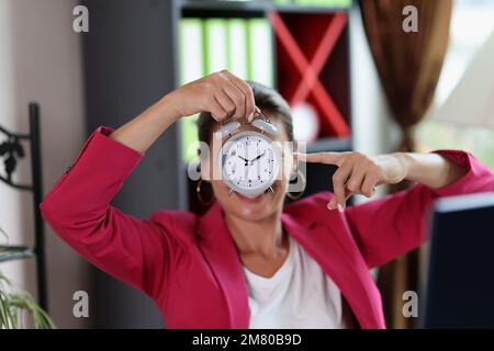 Die Frau im Büro zeigt auf den Wecker in ihrer Hand. Stockfoto