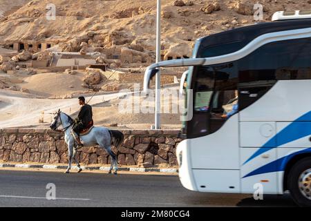 KONTRAST ZWISCHEN EINEM MANN AUF DEM PFERD UND EINEM TOURISTENBUS VOR DEM TAL DER ADLIGEN, WO DIE GRÄBER VIELER ADLIGER AUS DEM NEUEN REICH ZU FINDEN SIND, LUXOR, ÄGYPTEN, AFRIKA Stockfoto