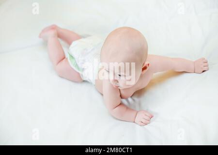 Ein kleines Kind lügt und erhebt seinen Kopf auf weißem Blut. Ein fröhliches Baby lernt, an den Griffen zu krabbeln und zu klettern. Kinderbetreuung in Windeln. Stockfoto