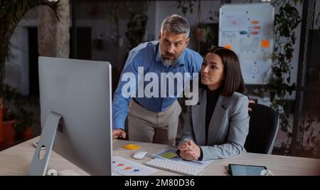 Kluge Geschäftsfrau, die mit dem leitenden Chef spricht, während sie im Büro auf den PC-Bildschirm schaut Stockfoto