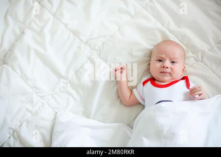 Ein winziges Neugeborenes auf einem riesigen Bett unter einer weißen Decke. Emotionen eines Neugeborenen. Ein kleines Mädchen hat ihre Mutter gesehen. Stockfoto