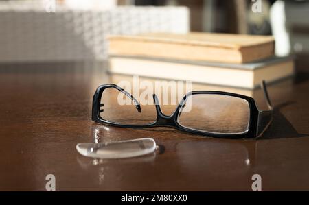 Ein zerbrochener Brillenrahmen liegt vor einem Stapel alter Bücher auf einem braunen Tisch Stockfoto