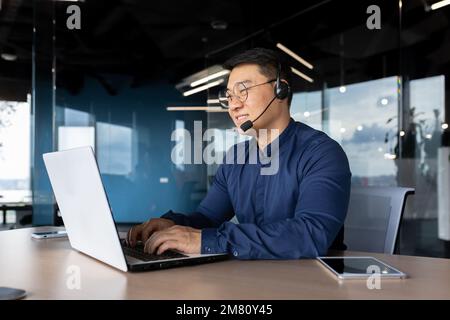 Ein junger männlicher asiatischer Lehrer hält Vorträge und nimmt die Prüfungen online per Laptop-Videoanruf ab. Sie sitzen im Büro am Tisch mit einem Headset und lächeln in die Kamera. Stockfoto