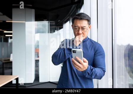 Ein besorgter und schockierter junger asiatischer Mann schaut in seine Hände. Liest Nachrichten, Nachrichten. Es steht im Büro, neben dem Fenster. Stockfoto