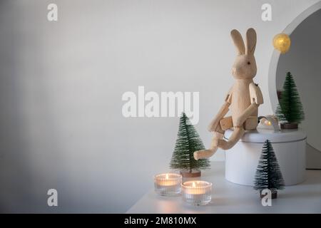 Ein schwenkbarer Holzhase, ein kleiner künstlicher grüner Weihnachtsbaum und beleuchtete Kerzen in Kerzenleuchtern auf einer weißen Kommode. Platz für Text. Christm Stockfoto