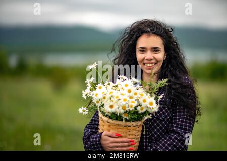 Eine Frau steht auf einem grünen Feld und hält einen Korb mit einem großen Strauß Gänseblümchen in den Händen. Im Hintergrund gibt es Berge und einen See. Stockfoto