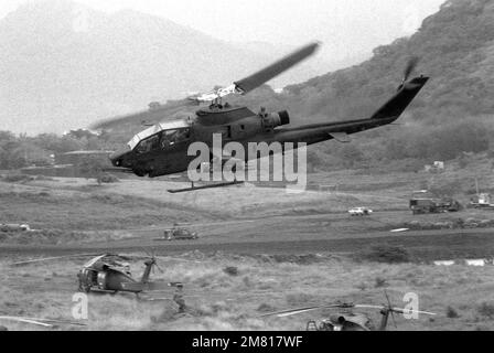 Ein AH-1 Cobra Helikopter fliegt über mehrere UH-60 Black Hawk (Blackhawk) Helikopter, die auf einem Feld am Point Salines Airfield während der multifunktionalen multinationalen Operation Urgent Fury geparkt sind. Betreff Betrieb/Serie: URGENT FURY Country: Grenada (GRD) Stockfoto