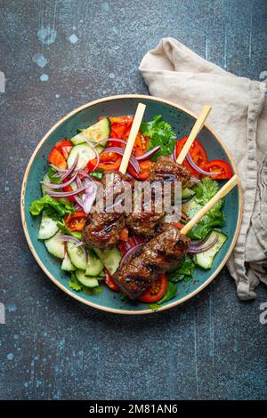 Gegrillte Spießfleisch-Rindfleisch-Kebabs auf Stöcken serviert mit frischem Gemüsesalat auf einem Teller auf rustikalem Betonhintergrund von oben. Traditioneller Mittelteil Stockfoto