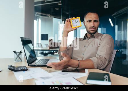 Unzufriedener hispanischer Mann, der in die Kamera schaut und die Uhr zeigt, Geschäftsmann, frustriert darauf zu warten, dass ein Kollege zu spät kommt, im Büro mit Dokumenten zu arbeiten, die ein Notebook im Büro verwenden. Stockfoto