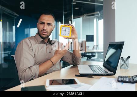 Unzufriedener hispanischer Mann, der in die Kamera schaut und die Uhr zeigt, Geschäftsmann, frustriert darauf zu warten, dass ein Kollege zu spät kommt, im Büro mit Dokumenten zu arbeiten, die ein Notebook im Büro verwenden. Stockfoto