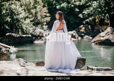 Eine wunderschöne Frau in einem langen weißen Kleid blickt in die Ferne auf einen wunderschönen See mit Blick auf die Schwäne von hinten Stockfoto