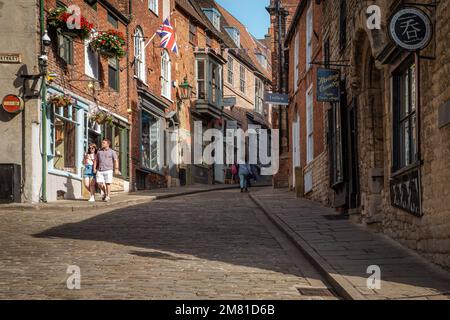 Einkäufer und Touristen genießen die malerischen Geschäfte, die eine steile Kopfsteinpflasterstraße säumen, auch bekannt als steiler Hügel in Lincoln. Stockfoto