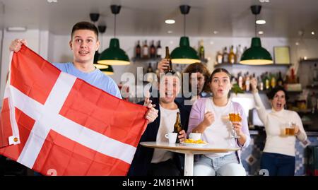Brüllende junge Sportfans, die auf ihre Lieblingsmannschaft feuern und mit der Flagge Dänemarks wedeln, während sie gemeinsam im Pub das Spiel sehen Stockfoto