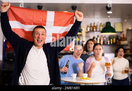 Begleitung junger begeisterter Erwachsener-Fans, die mit der Flagge Dänemarks wedeln und die Nationalmannschaft mit Bier im Pub unterstützen Stockfoto