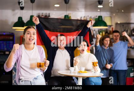 Gesellschaft von schreienden jungen erwachsenen Sportfans, die die Flagge Deutschlands schwenken und die Nationalmannschaft mit Bier im Pub unterstützen Stockfoto