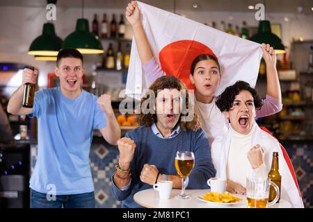 Begeisterte Gesellschaft von jungen Sportfans, die die Flagge Japans schwenken und die Lieblingsmannschaft mit Bier im Pub unterstützen Stockfoto