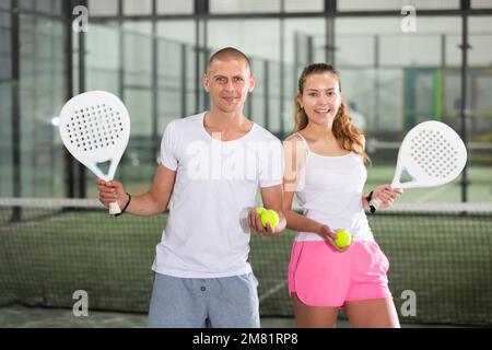 Ein paar fröhliche junge Paddle-Tennisspieler stehen auf dem Hallenplatz Stockfoto