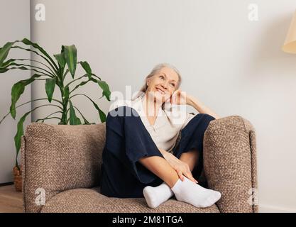 Fröhliche Seniorin, die sich zu Hause in einem Sessel entspannt. Lächelnde alte Frau in der Freizeit im Wohnzimmer. Stockfoto