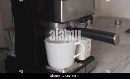 Nahaufnahme von heißem Kaffee, der aus dem Kaffeeauslauf gegossen wird, Zubereitung von Morgenkaffee mit der Kaffeemaschine. Hochwertiges Foto Stockfoto