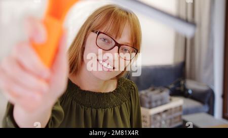 Glückliche Frau mit Brille und Ingwerhaar, die die Kamera mit einem Wischer reinigt, Hausfrauenkonzept. Hochwertiges Foto Stockfoto