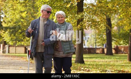 Seniorin, die ihrem blinden Mann hilft, durch den Park zu gehen. Hochwertiges Foto Stockfoto