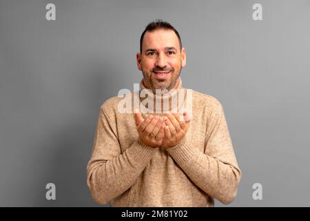 ein bärtiger latino in einem Rollkragenpullover, der etwas Fantasielles in den Händen nahe der Brust bietet, isoliert auf grauem Studiohintergrund Stockfoto