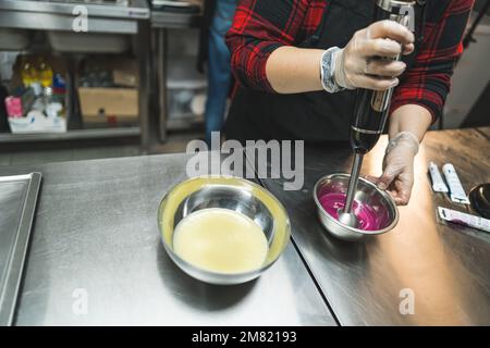 Eine unbekannte Konditorin bereitet ein rosafarbenes Sahnehäubchen für einen Kuchen zu und peitscht ihn mit einem Mixer in einer Edelstahlschüssel. Hochwertiges Foto Stockfoto