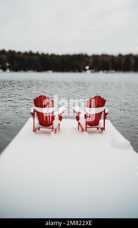 Rote Adirondack muskoka Stühle am Ende des verschneiten Docks im Winter. Stockfoto