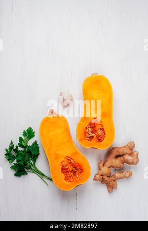 Roher Butternusskürbis mit Ingwer, Knoblauch und Petersilie auf einem weißen Holztisch. Zutaten für Kürbissuppe. Draufsicht, flach liegend. Stockfoto