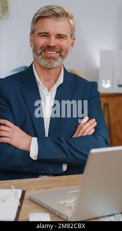 Selbstbewusster, glücklicher, erwachsener Geschäftsmann, der mit einem Lächeln in die Kamera schaut, am Arbeitsplatz des Heimbüros. Gesichtsbild eines grauen Freiberufler-Maklers mittleren Alters, Unternehmer, Firmenführer-Boss Stockfoto