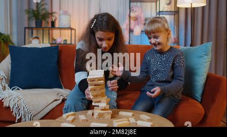 Teenager und kleine Schwestermädchen bauen einen Turm aus hölzernen Ziegeln und verlieren den Brettspielwettbewerb. Geschwister, Kinder oder beste Freunde, die Spaß mit dem Turm auf dem Tisch haben und zu Hause mit Bausteinen spielen Stockfoto